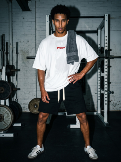 Man in a gym wearing black heavyweight raw-edge shorts, a white Pheuw tee and a towel over his shoulder